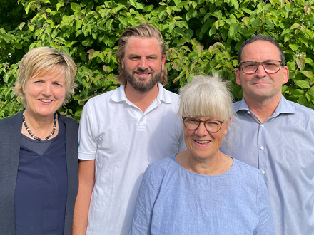 Auf dem Bild sind vier Personen vor einer grünen Hecke zu sehen.