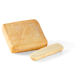 Auf dem Bild ist ein Stück Käse zu sehen, das in Quaderform mit einer aufgeritzten Oberfläche sowie einer abgeschobenen Käsescheibe daneben präsentiert wird.