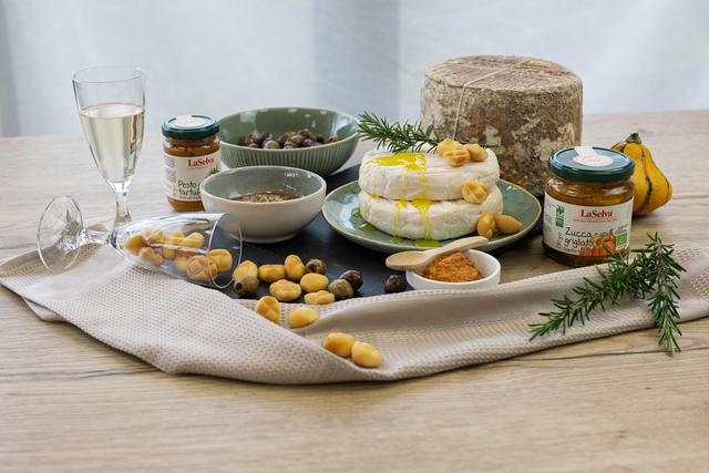 Das Bild zeigt eine dekorative Anrichtung mit verschiedenen Lebensmitteln, darunter Käse, Oliven, Pesto, eine Kürbismarmelade, ein paar Snacks und Gläser mit Getränken.